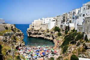 Crowded beach with colorful umbrellas in Polignano a Mare, Italy, with scenic cliffside buildings.