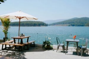 Enjoy a serene lakeside dining experience with wooden tables under a sun umbrella.