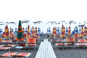 Colorful beach umbrellas and chairs beside the sea with boats in the background, offering a serene summertime vibe.
