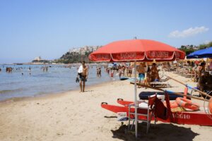Crowded beach with red lifeguard station and swimmers under a clear blue sky.